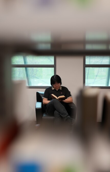 Photo of a person reading a book, with the camera positioned to capture the shot through a shelf of books in a library.