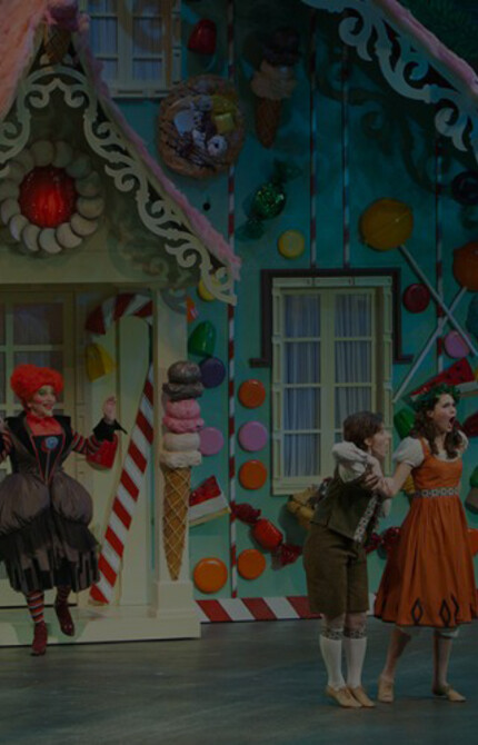 Photo of a scene from Hansel and Gretel, with the characters Hansel, Gretel, and the Witch standing in front of the candy house