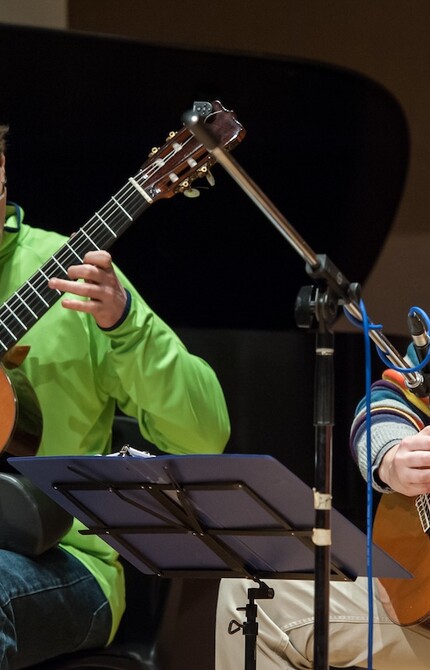 Two guitarists performing, surrounded by performance equipment, both focused on the music on a stand.