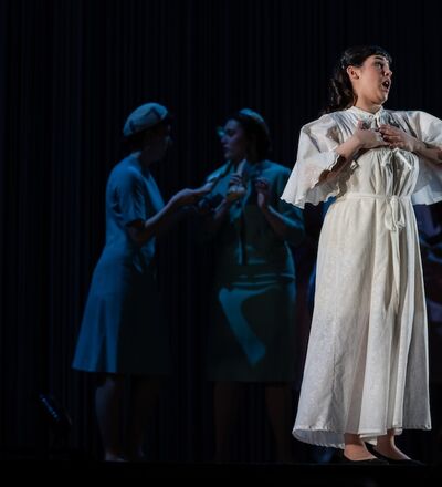 Scene from an opera, with a person singing in a white dress, while others are conversing in the background.