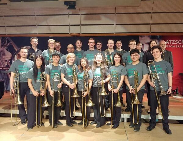 Group of people in grey shirts standing together, each holding a trombone