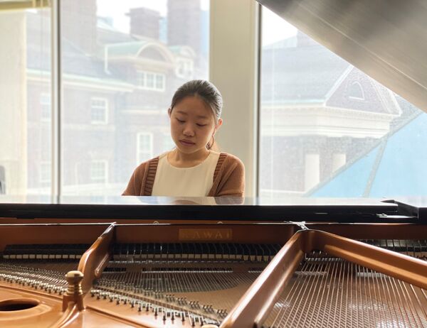"Person wearing a cardigan playing a grand piano, positioned in front of large windows that display a building in the background, with natural light illuminating the scene.