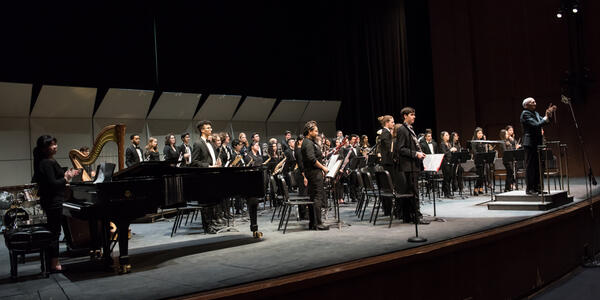 Large ensemble bowing to the audience on a larger stage, illuminated by stage lights, showing appreciation after their performance.