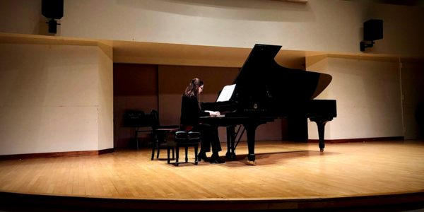 Person playing grand piano on the Walter Hall stage, with stage lighting.
