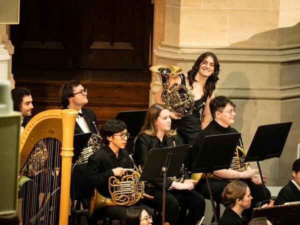 French horn soloist standing in the French horn section of an ensemble, wearing performance attire, surrounded by other musicians.