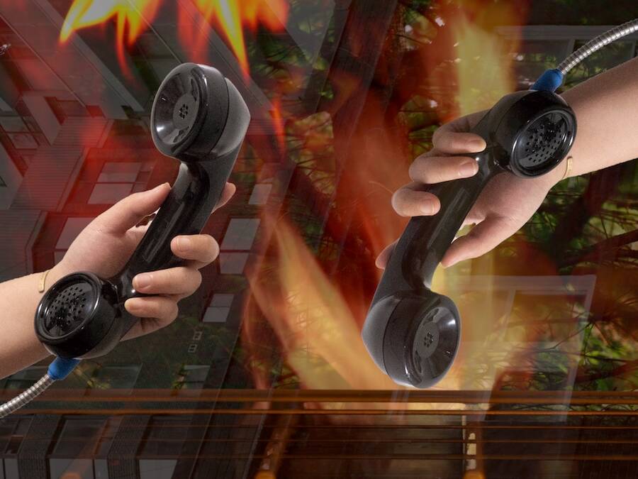 Two hands Holding telephones in front of an edited backdrop of of buildings, trees and fire