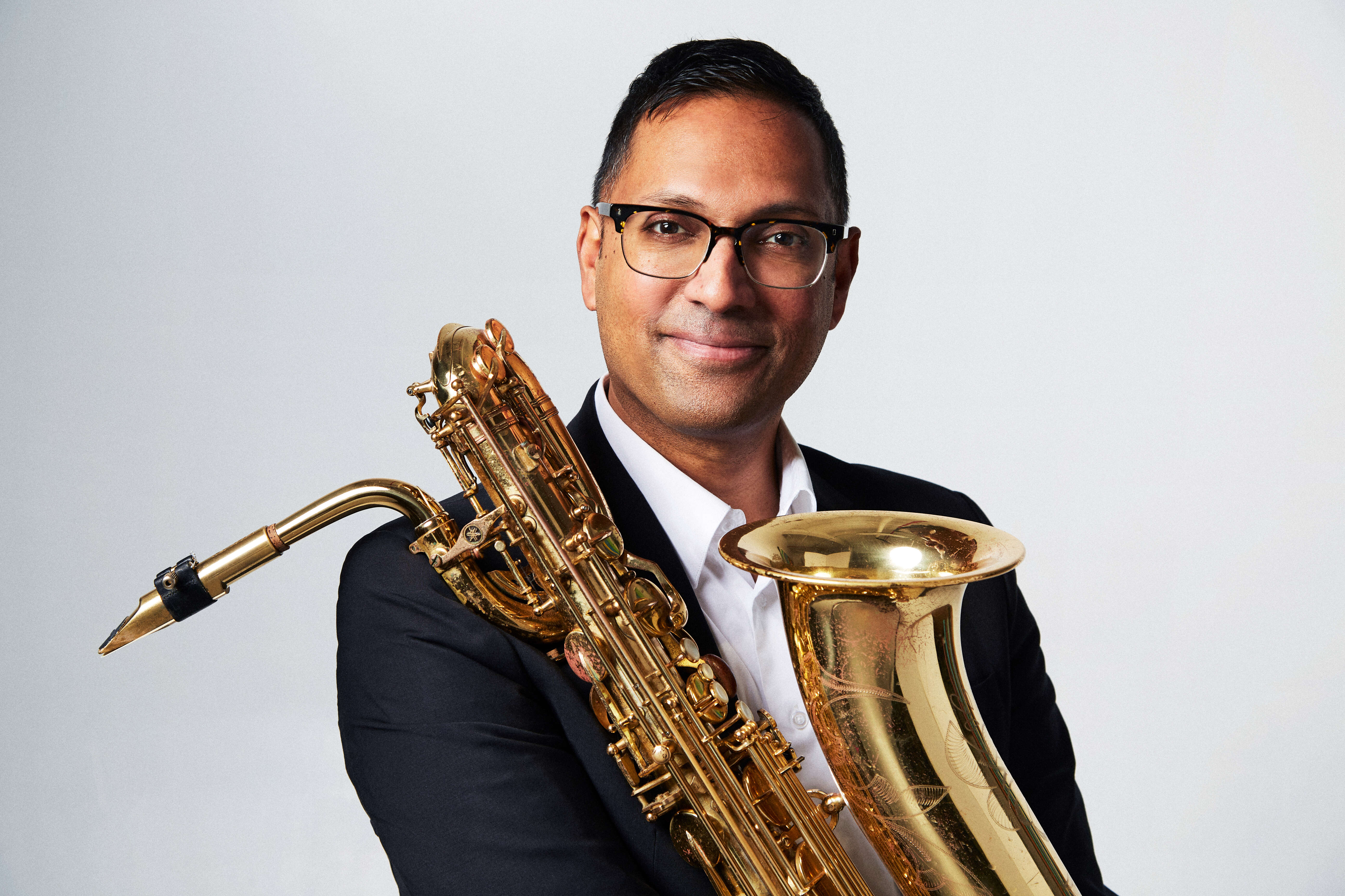 Professional headshot portrait of Shirantha Beddage, smiling and holding his saxophone in front of him against a white backdrop.