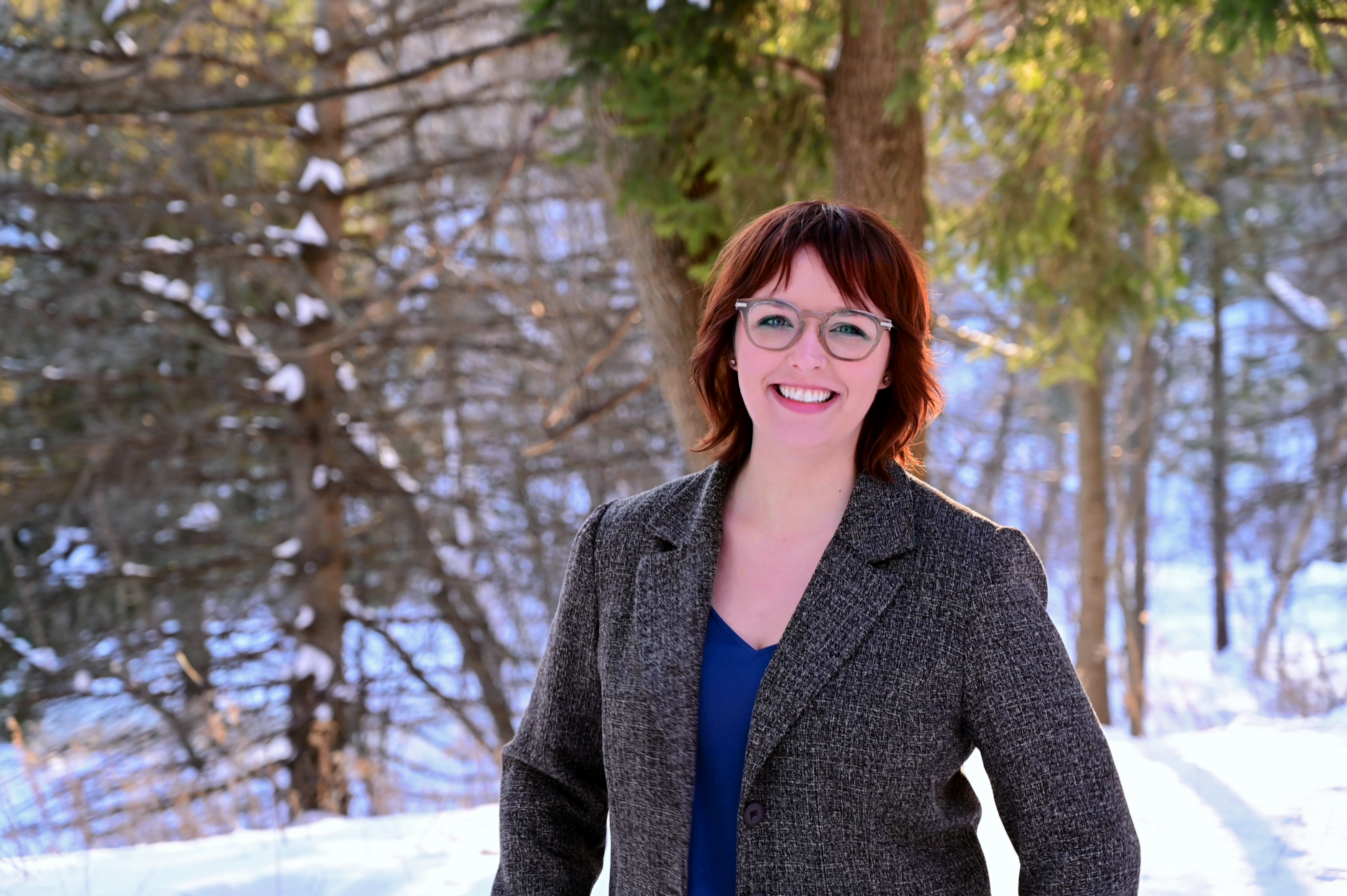Portrait of Avery Brzobohaty standing outdoors, in front of a wooded area with a grey jacket on