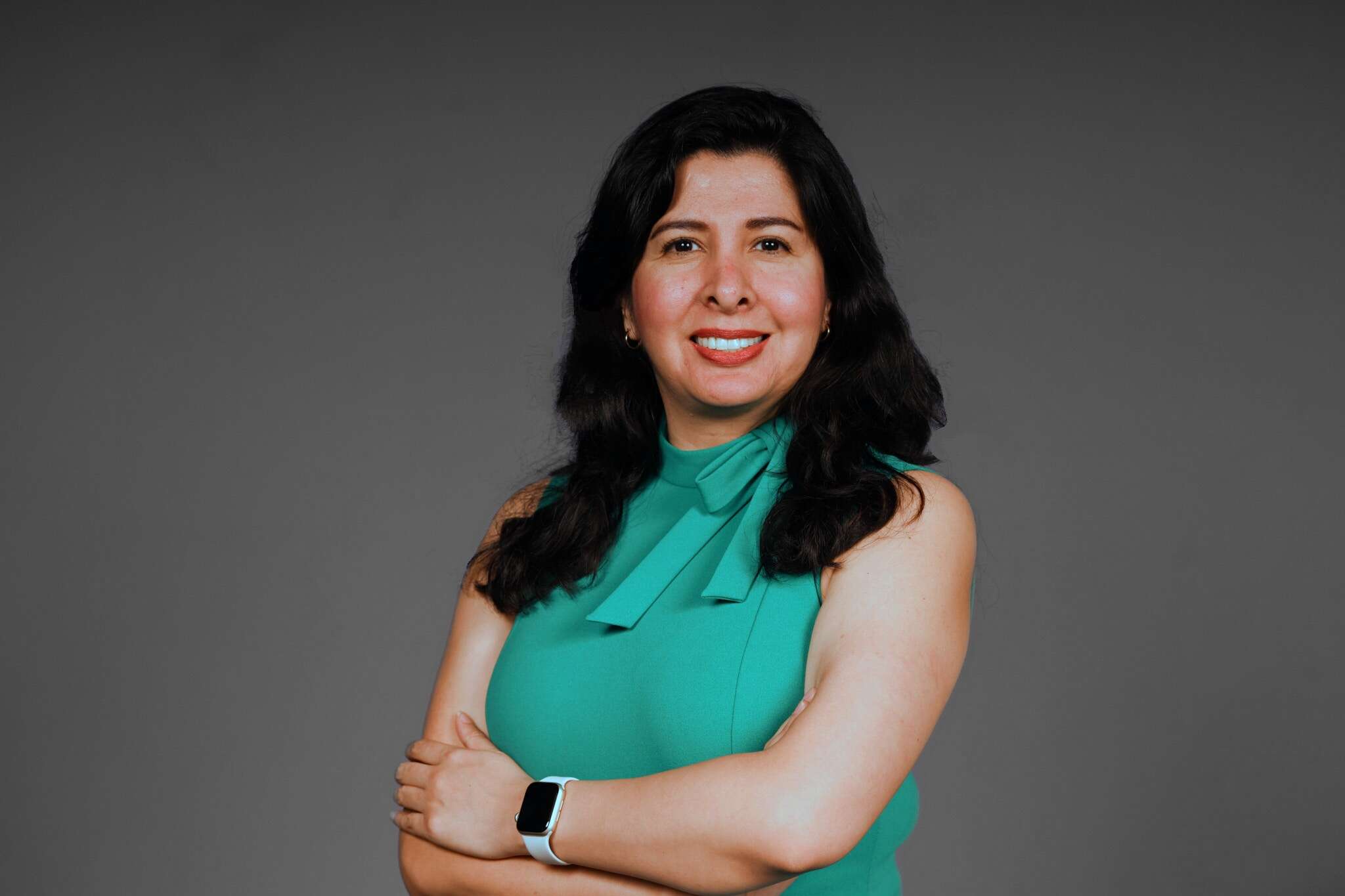 Professional headshot of Cristina Huerta, standing against a dark grey backdrop with her arms crossed and wearing a turquoise top.