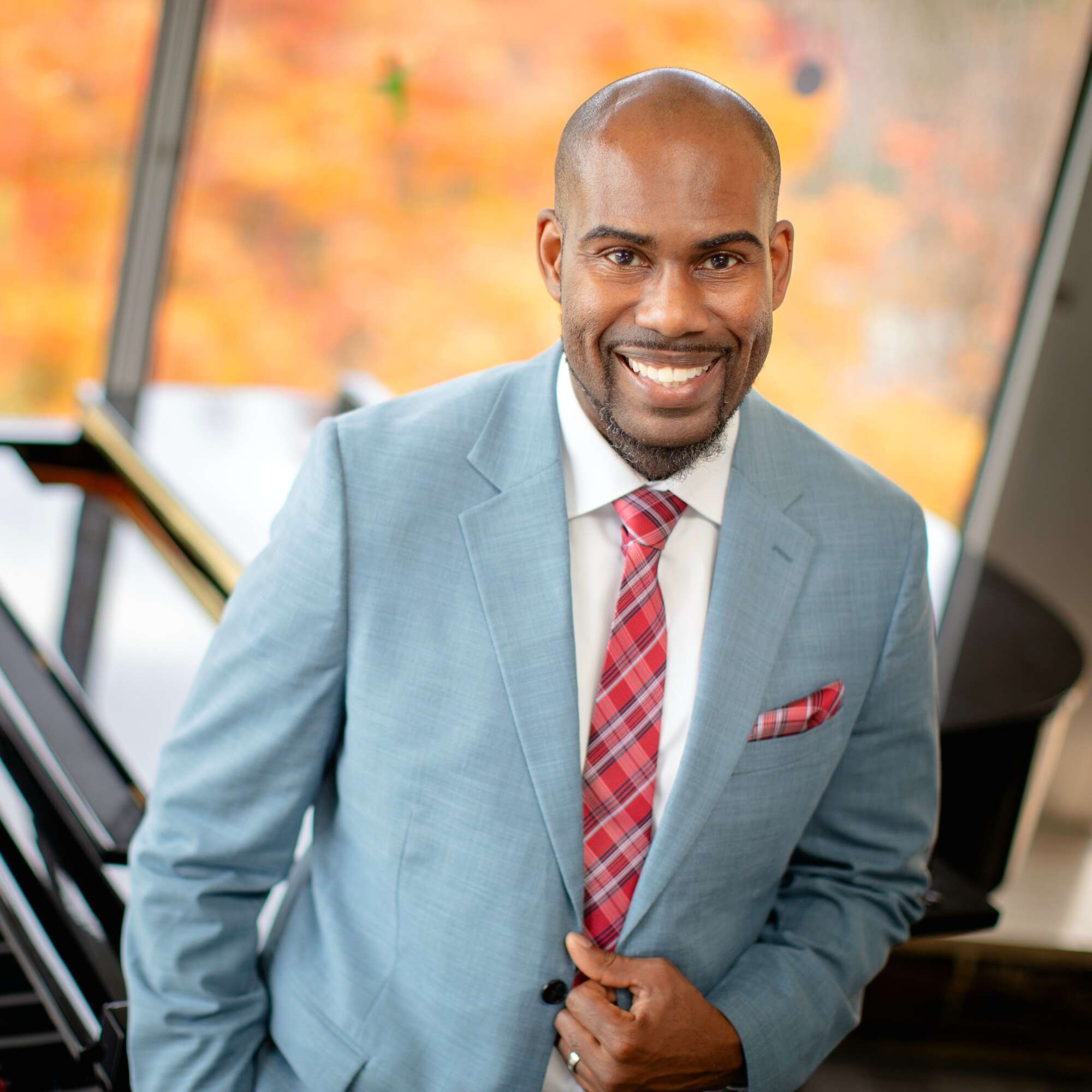 Portrait of a man smiling in front of a piano next to a window, wearing a grey suit with a red plaid tie.