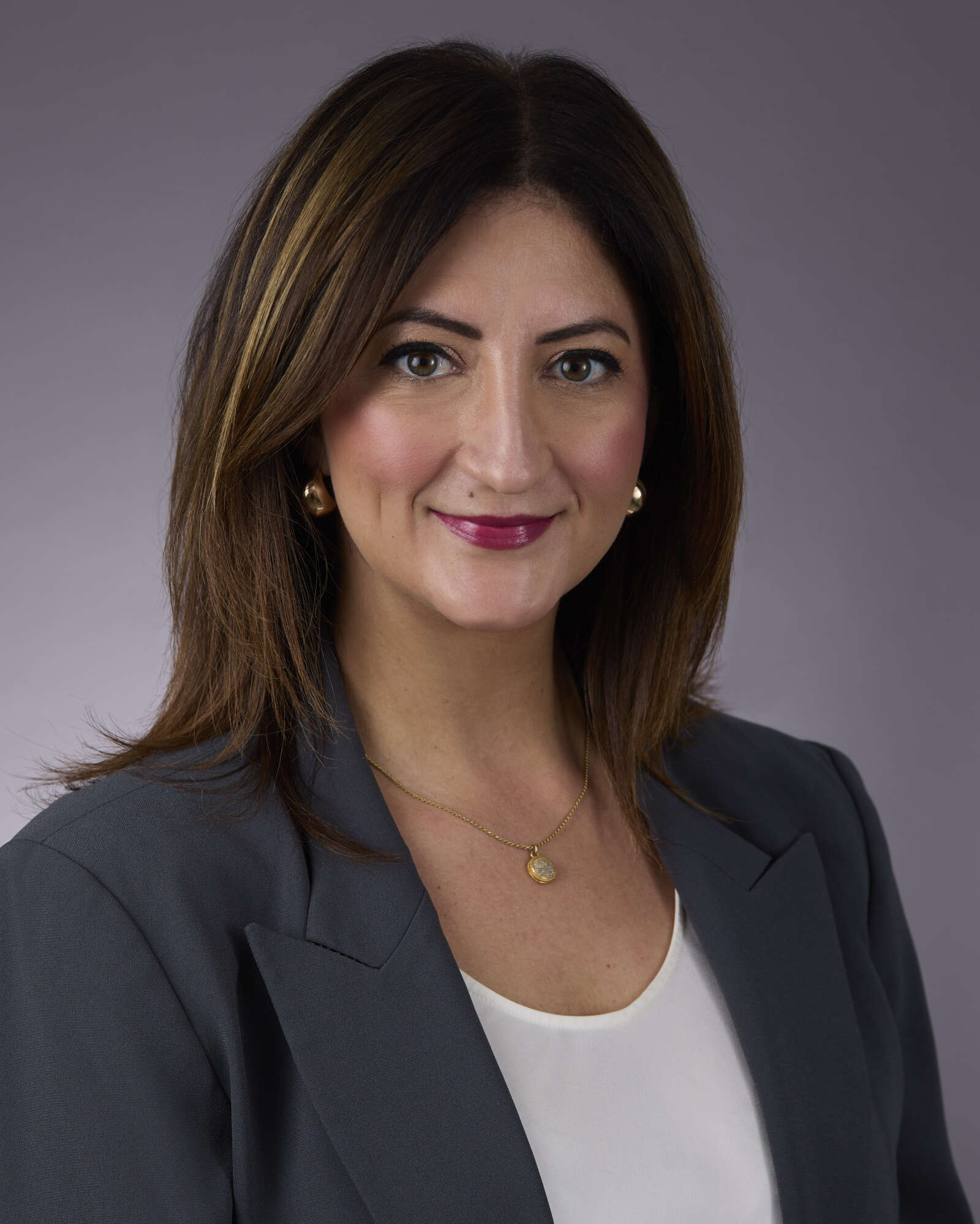 Portrait of woman with brown hair smiling, wearing dark blazer and white top, against a dark grey backdrop