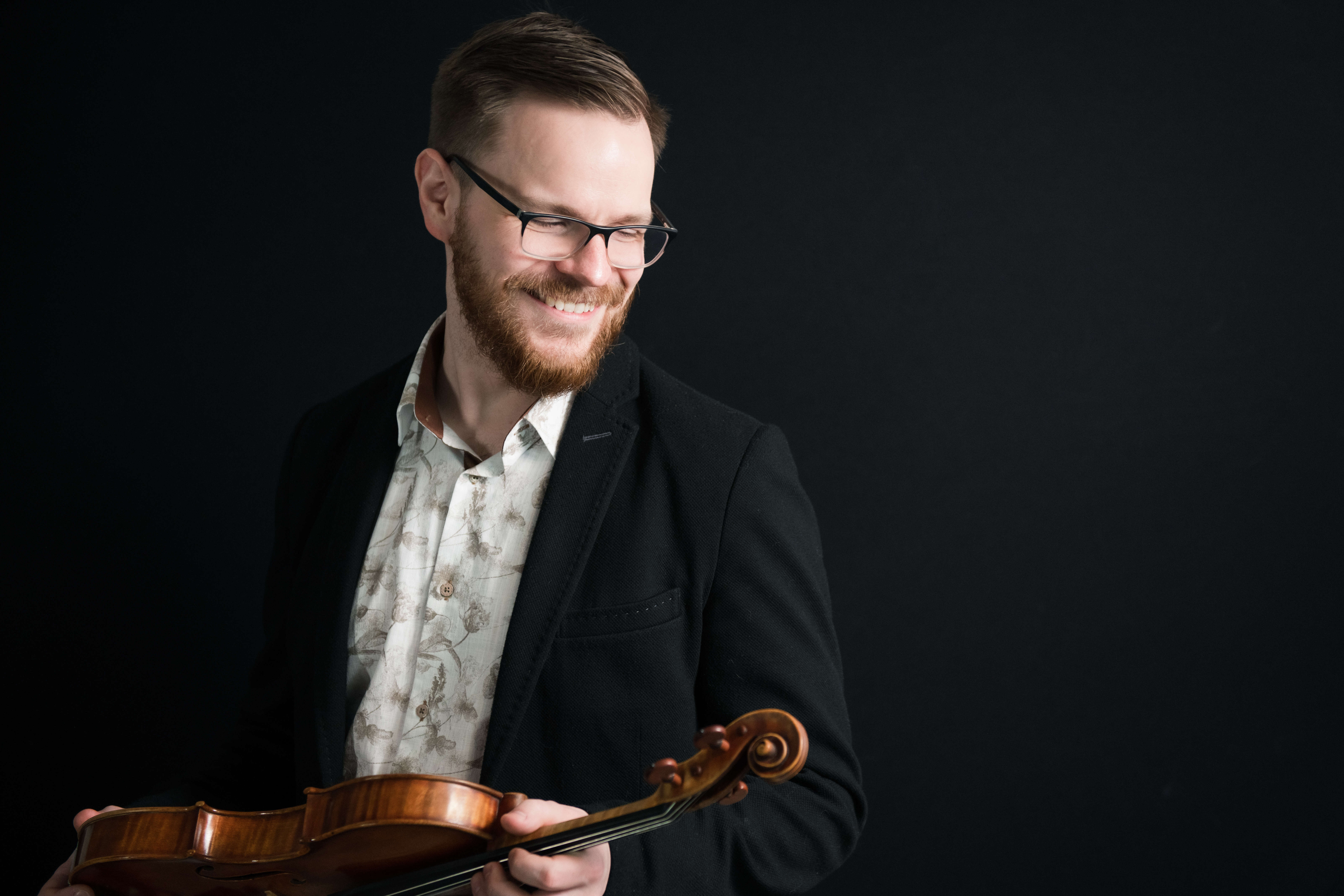 Person smiling, wearing glasses, a button-down shirt, and a blazer, holding a violin.