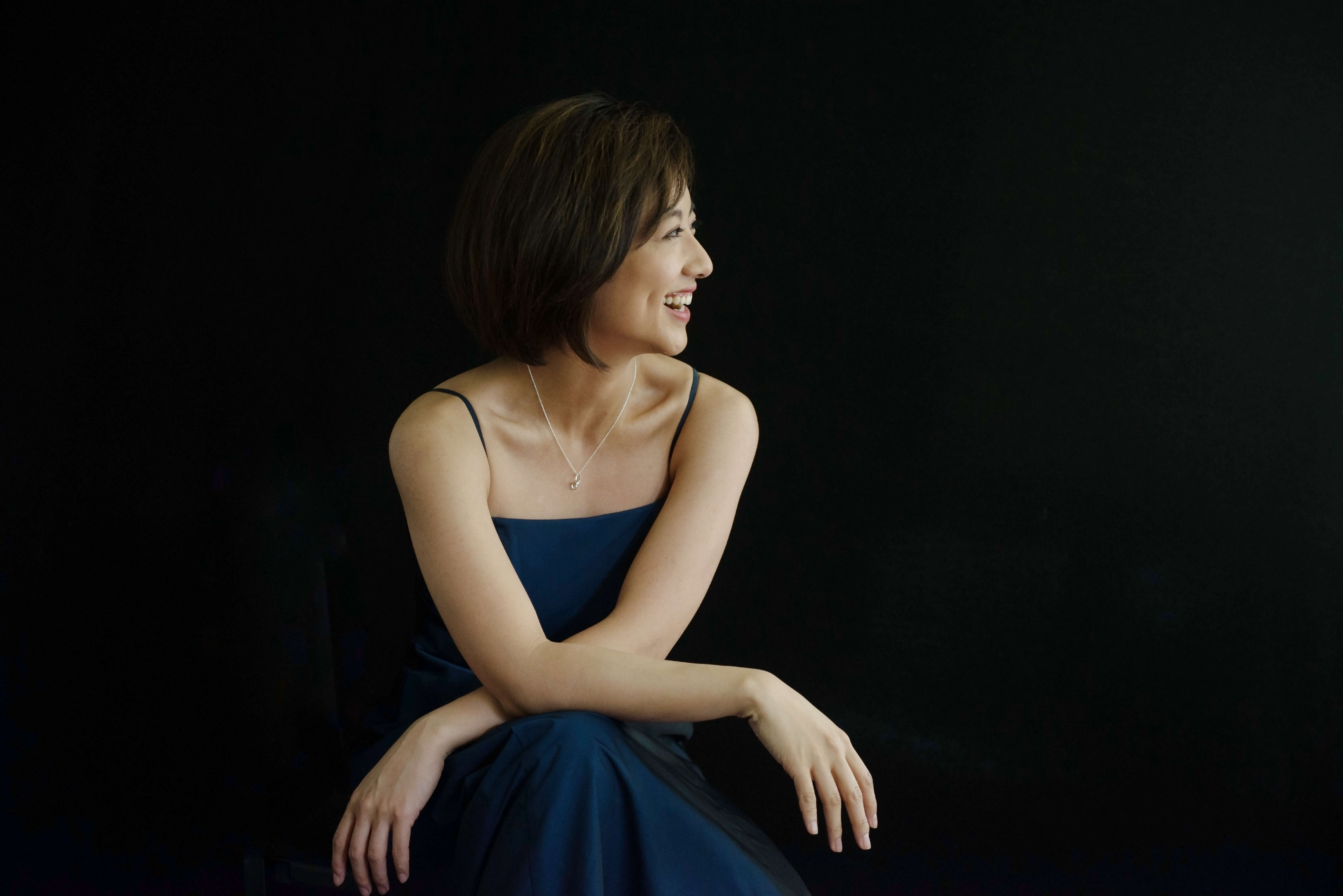 Person with short hair, wearing a blue dress, posed in front of a dark background.
