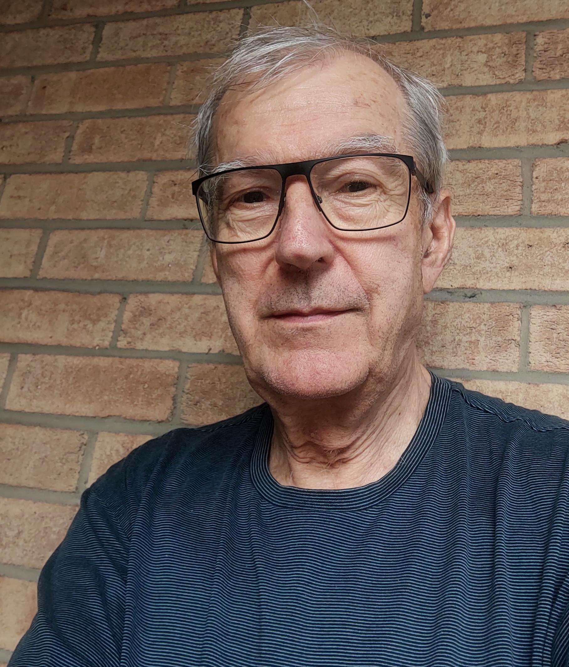 Headshot of an older person wearing a blue striped shirt and glasses, standing in front of a brick wall.