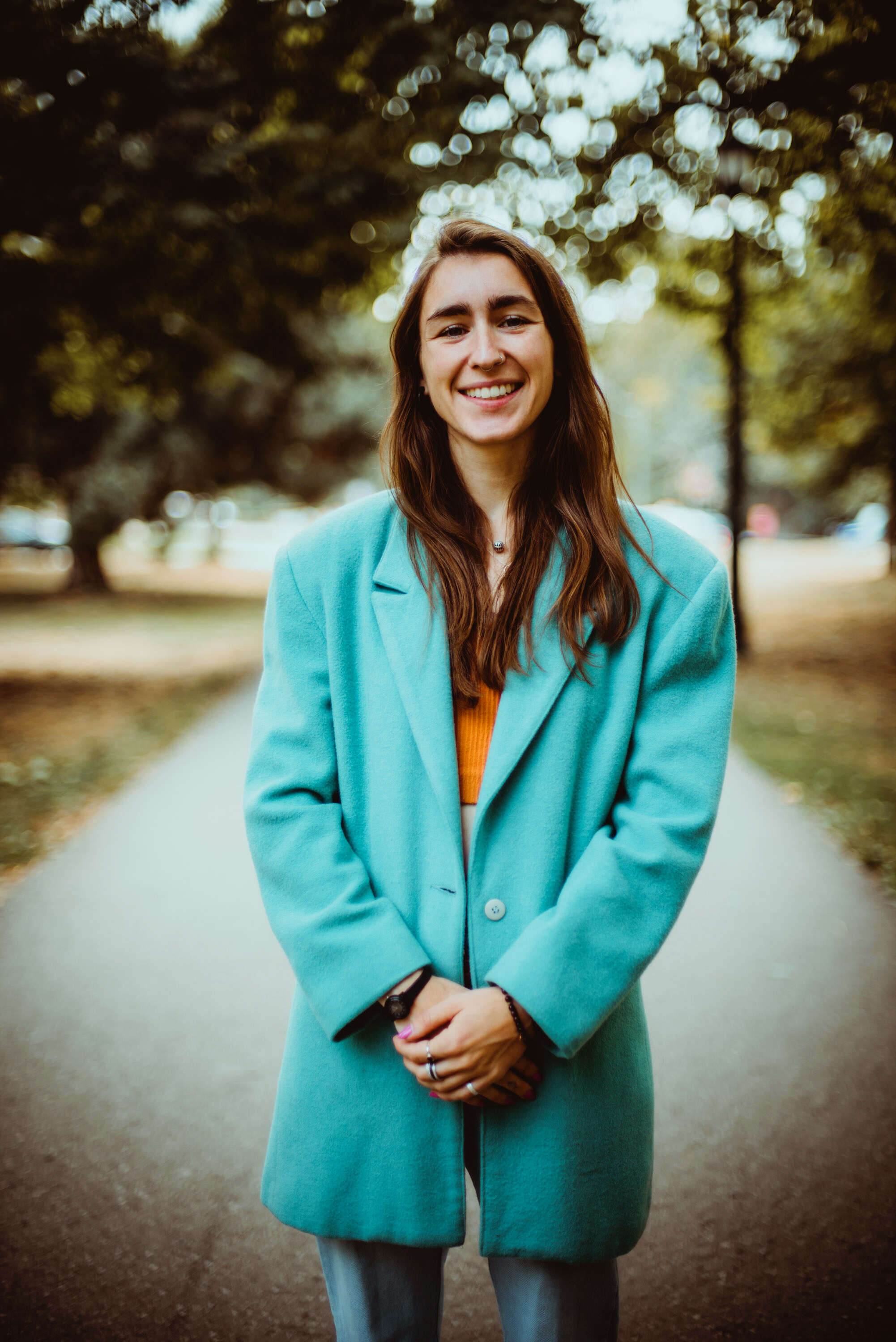 Person with long hair, wearing a teal blazer, smiling while standing in the middle of a wooded path.