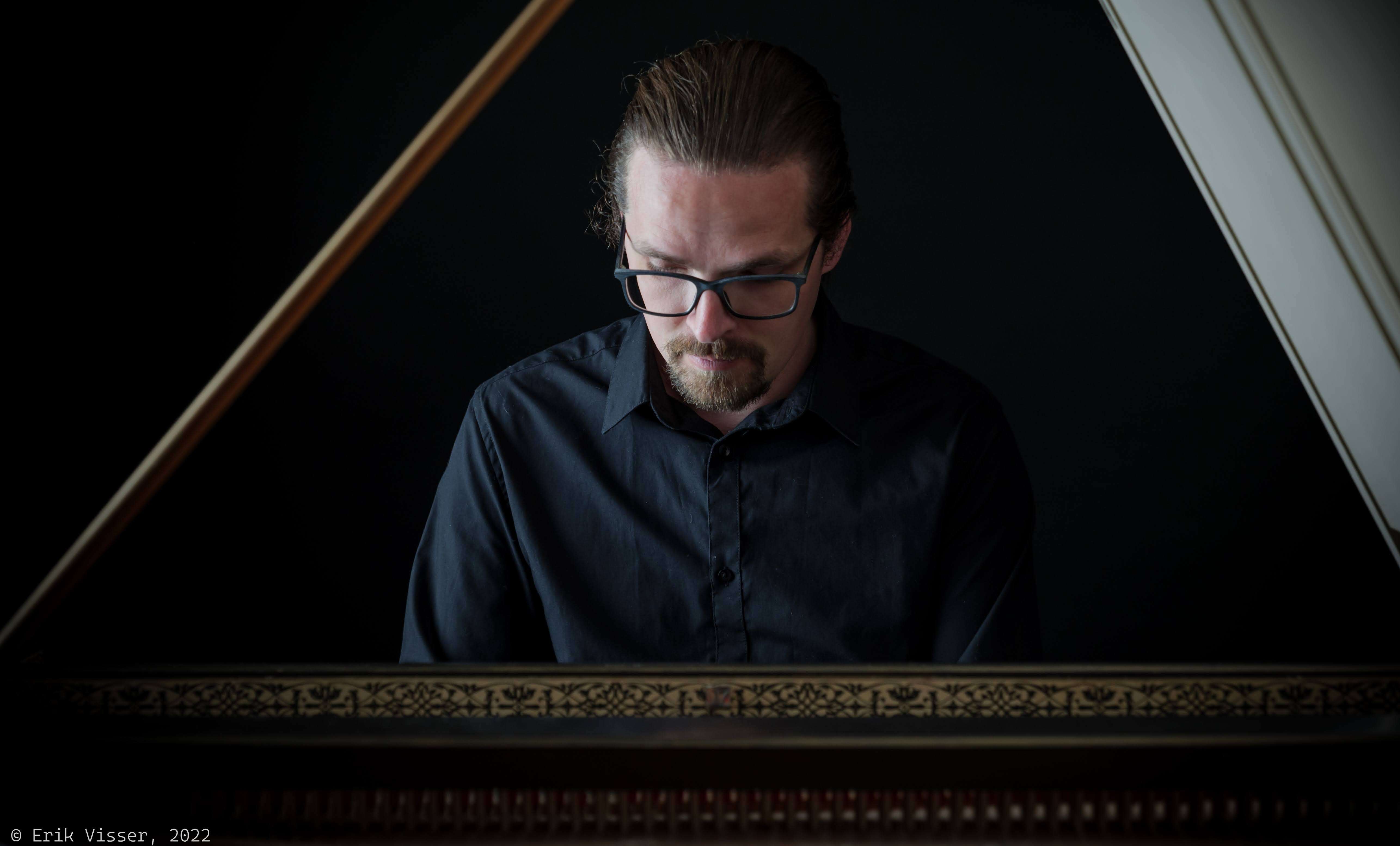 Person with longer hair wearing glasses and a black button-down shirt, playing the piano.