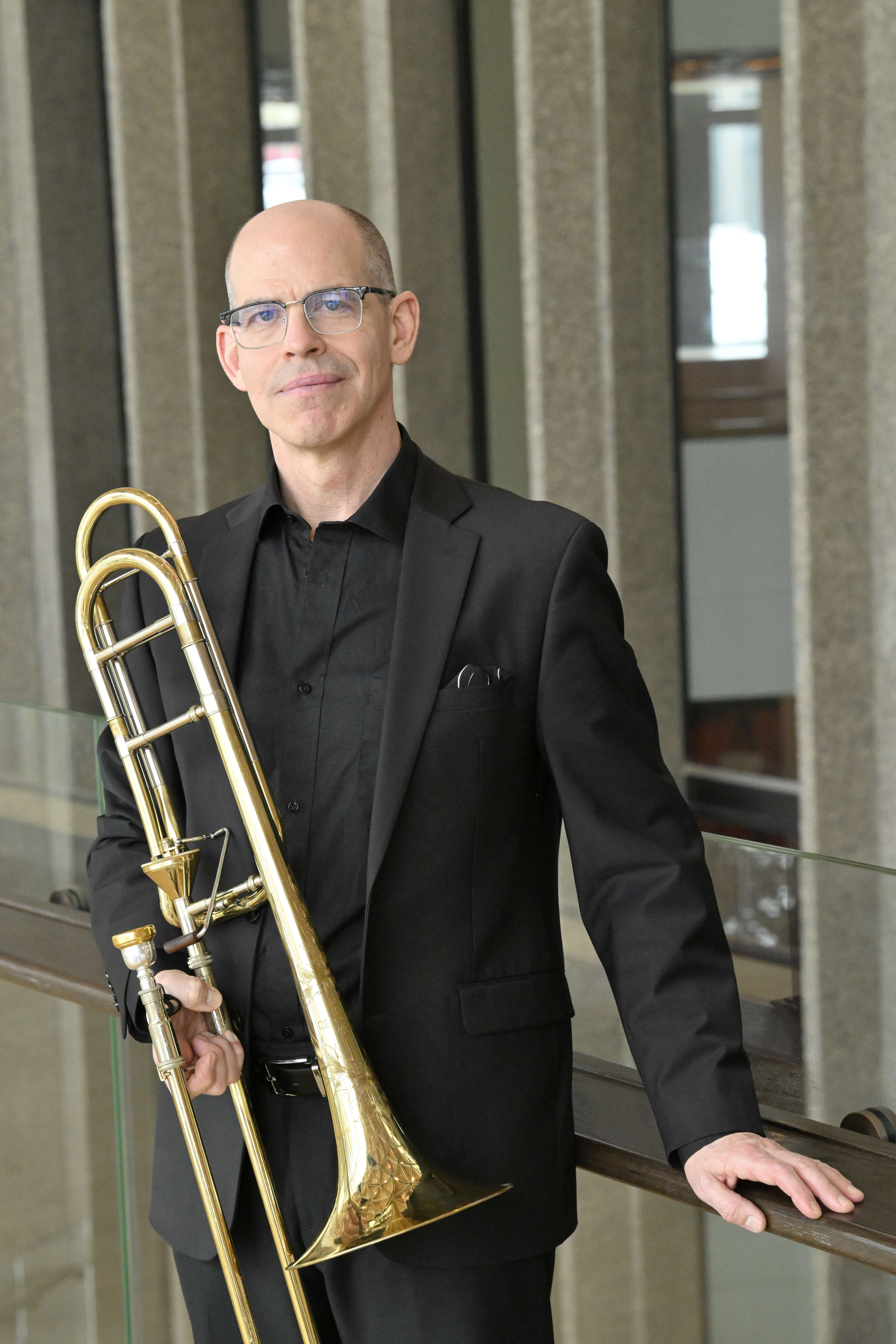 Photo of a man in a suit and glasses, holding a trombone, with windows in the background.