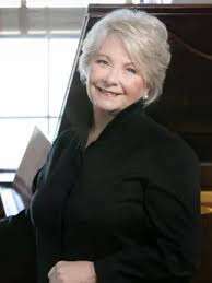 Older woman with a short haircut, smiling, wearing a black blouse, posed in front of a piano.