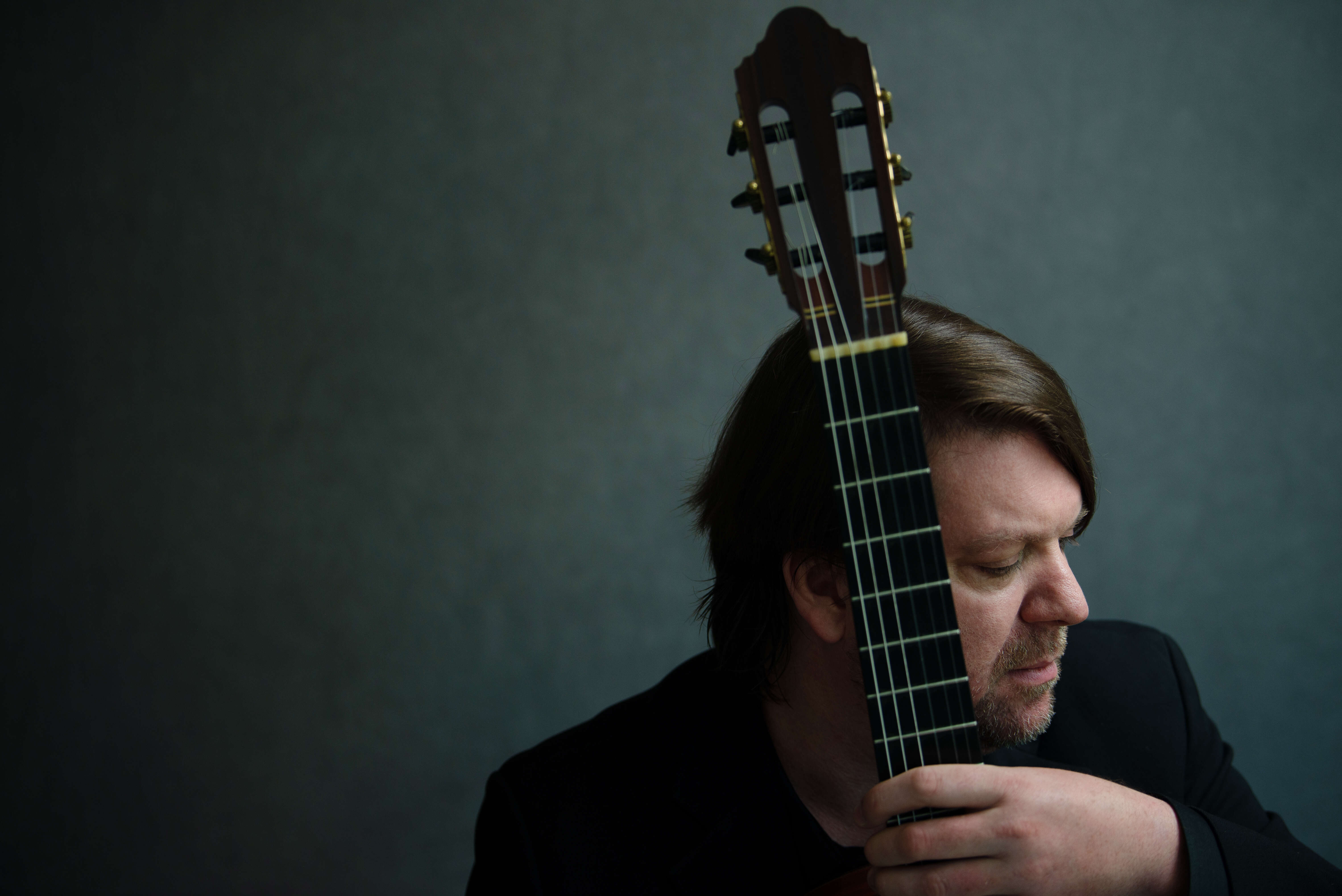 Person wearing a black jacket holding a guitar. The photo focuses on their profile, with the fingerboard of the guitar partially obscuring it.