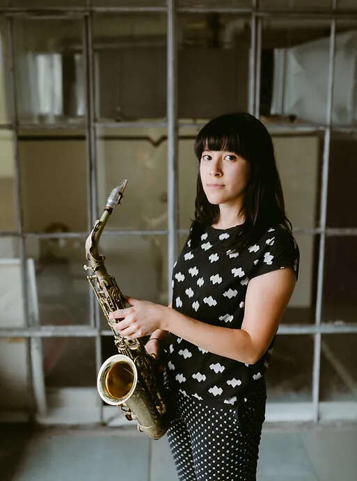 Person with bangs wearing a polka dot outfit, holding a saxophone while standing in an abandoned building.