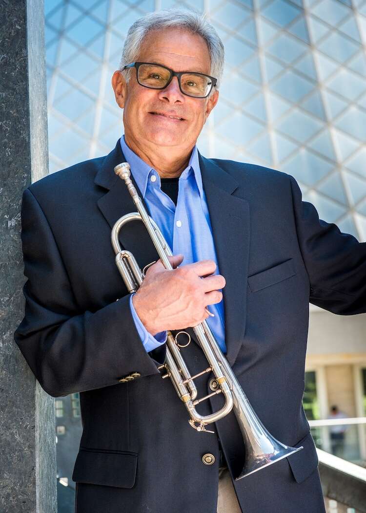 Person wearing a suit, holding a trumpet in front of a glass building.