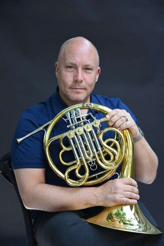 Headshot of a person with no hair in a blue polo shirt sitting in a chair holding a French horn with a dark grey background.