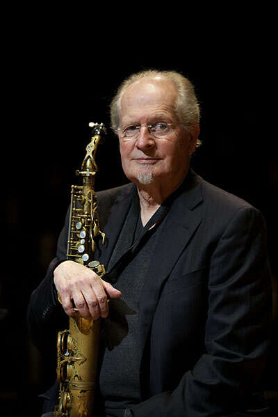Headshot of a person wearing glasses and a suit, holding a saxophone.