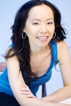 A headshot of a person with long black hair and hoop earrings, wearing a black tank top, leaning on a table.