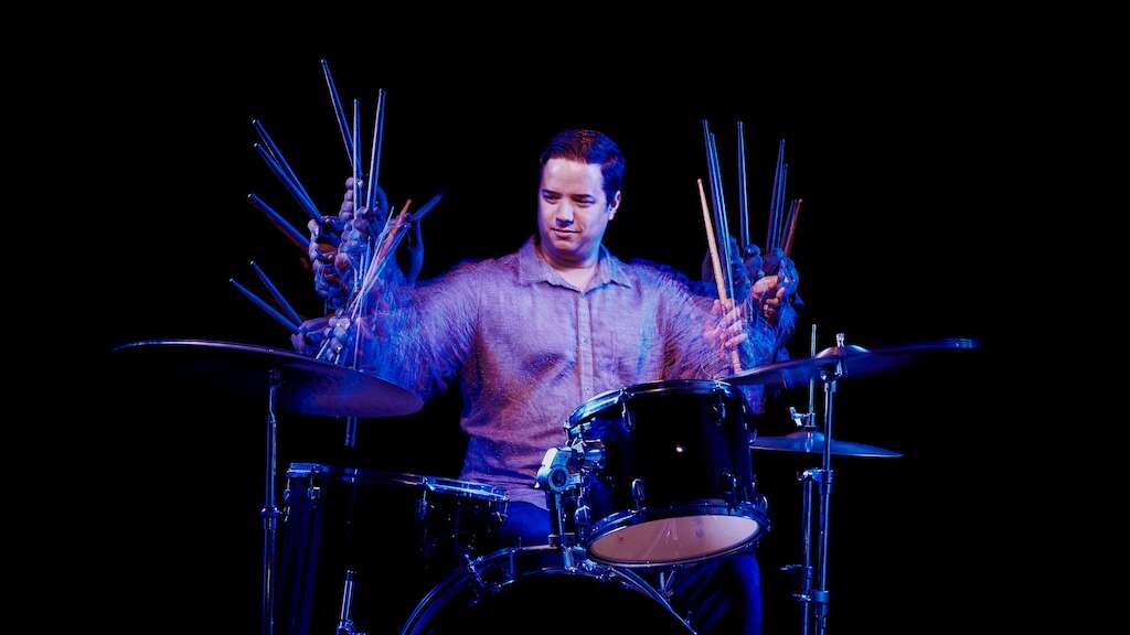 A person wearing a grey button-up shirt plays a drumset under blue and purple lighting, set against a black background.