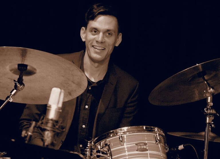 Black and white photo of a person wearing a button-up shirt and suit jacket, smiling while sitting in front of a drum set with a microphone.