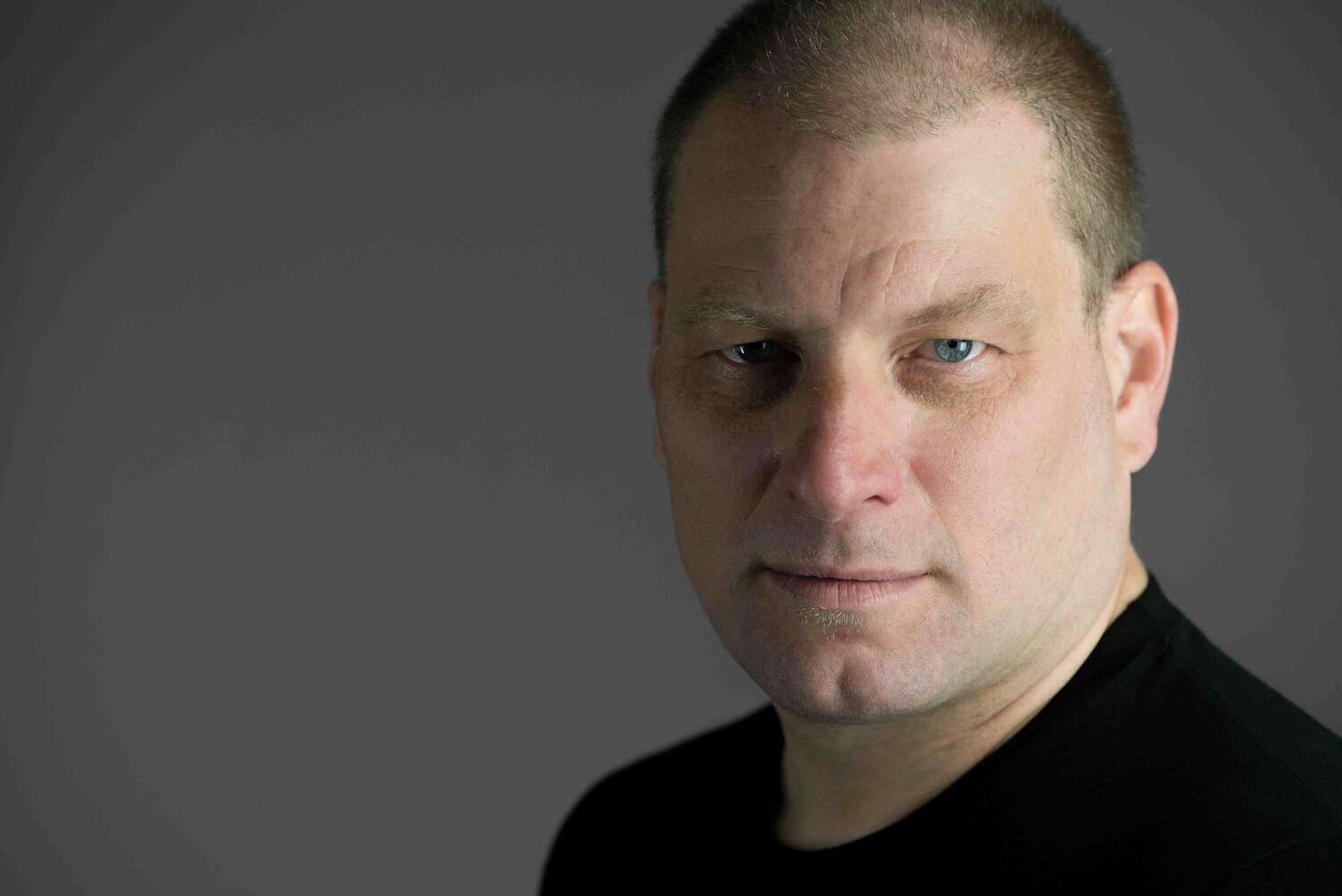 Headshot of a person wearing a black t-shirt in front of a grey background with a serious expression.