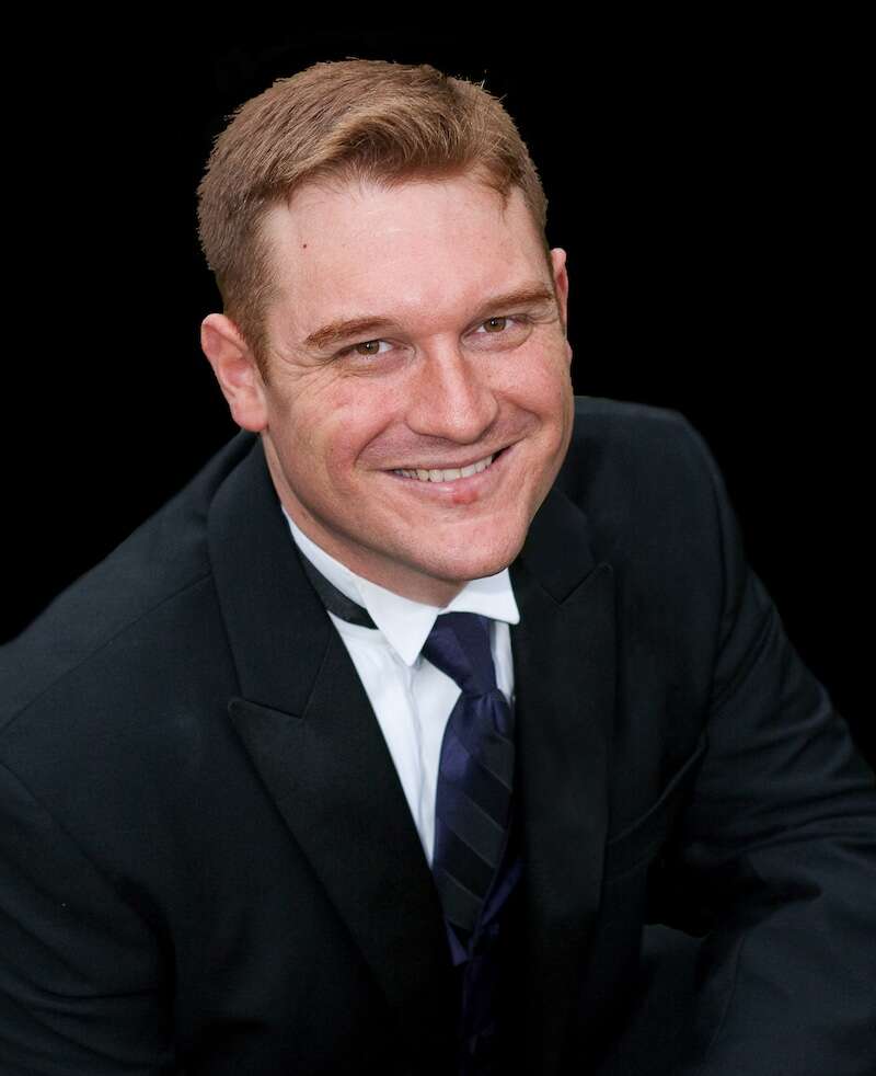 A headshot of a person wearing a suit, smiling and looking up at the camera, set against a black background.