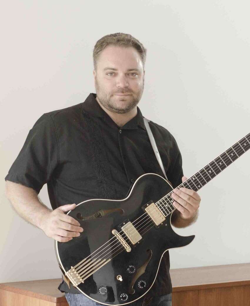 A person wearing a black short-sleeve button-up shirt is holding a guitar, leaning against a wooden cabinet in front of a white wall."