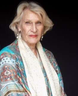 Headshot of a person wearing a colorful blouse and white pagmenia, looking at the camera with a confident expression against a dark background.