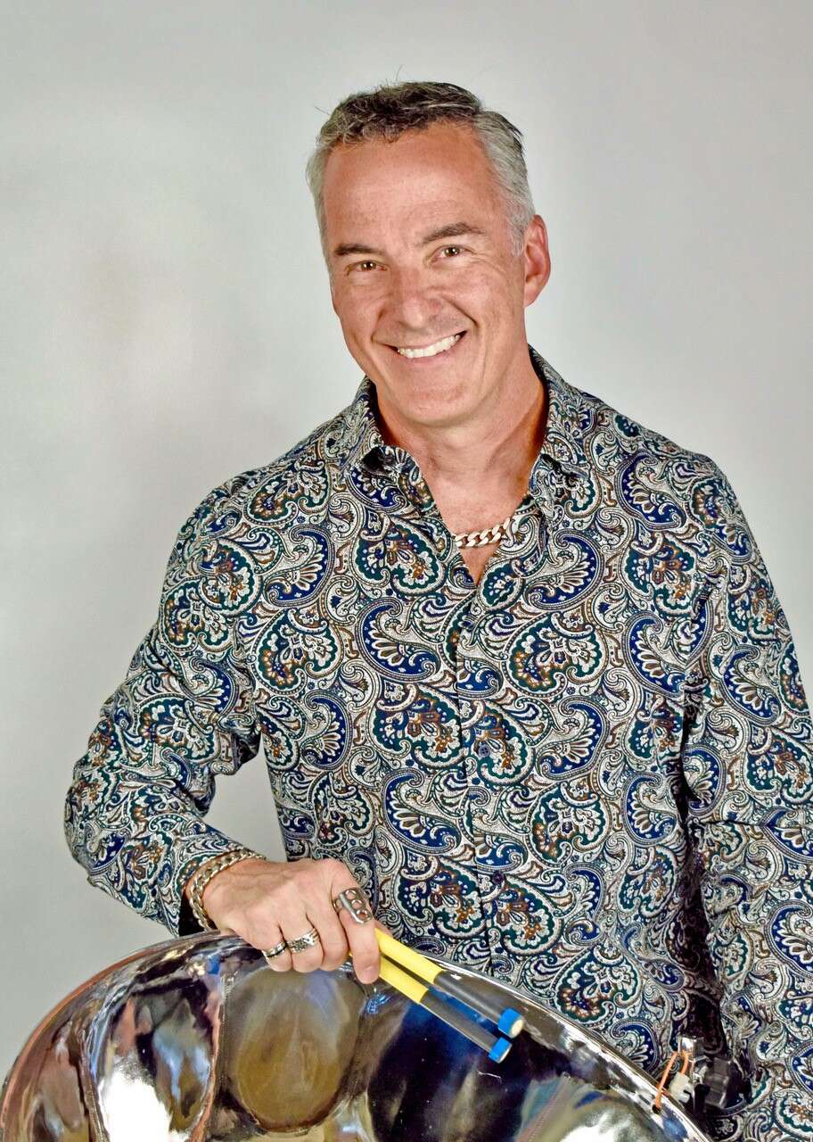 person smiles while wearing a vibrant blue and green patterned shirt, holding a steel pan drum and mallets, set against a grey background.