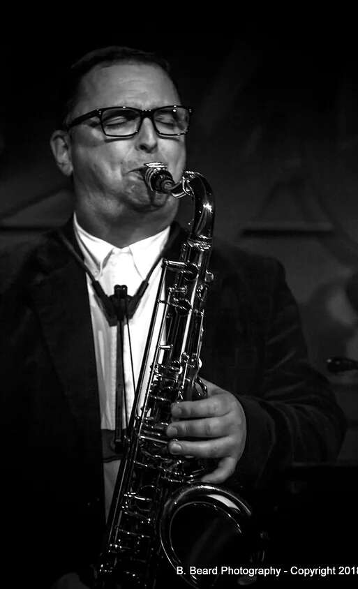 Black and white photo of a person wearing a suit and glasses, playing the tenor saxophone, captured in a moment of focused performance.
