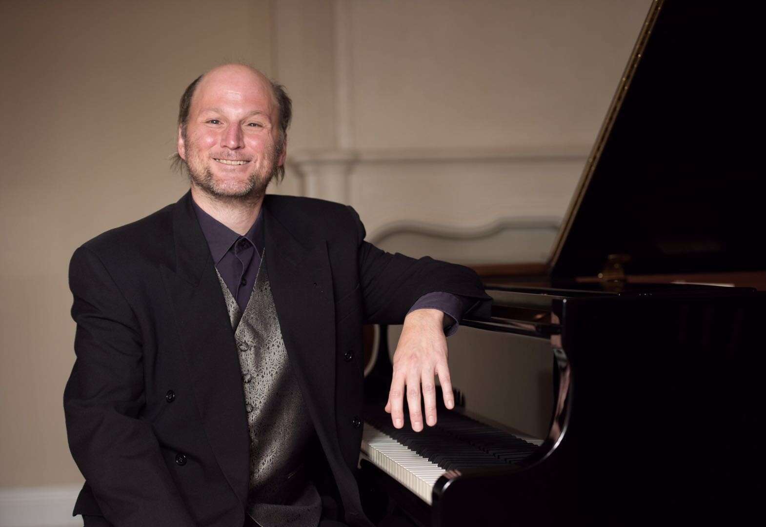 Person wearing a suit, smiling, and leaning on a grand piano.