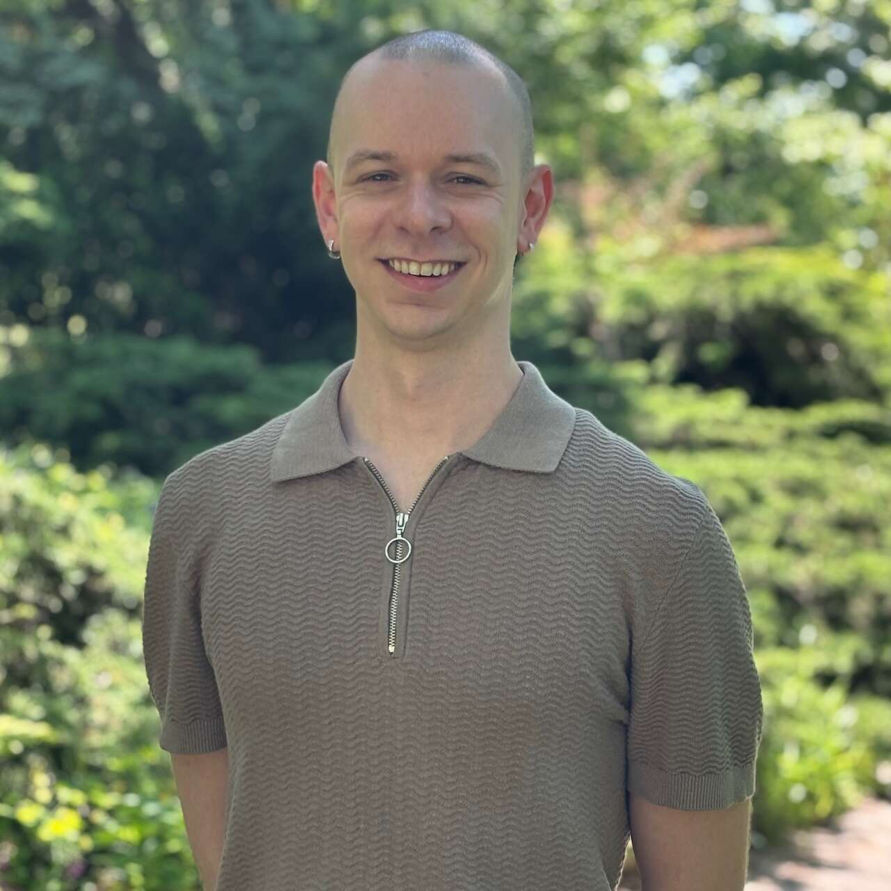 Person with buzz cut wearing in brown shirt. Blurred greenery in the background