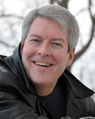 A headshot of a person with short grey hair smiling, wearing a leather jacket, standing in front of a tree on an overcast day