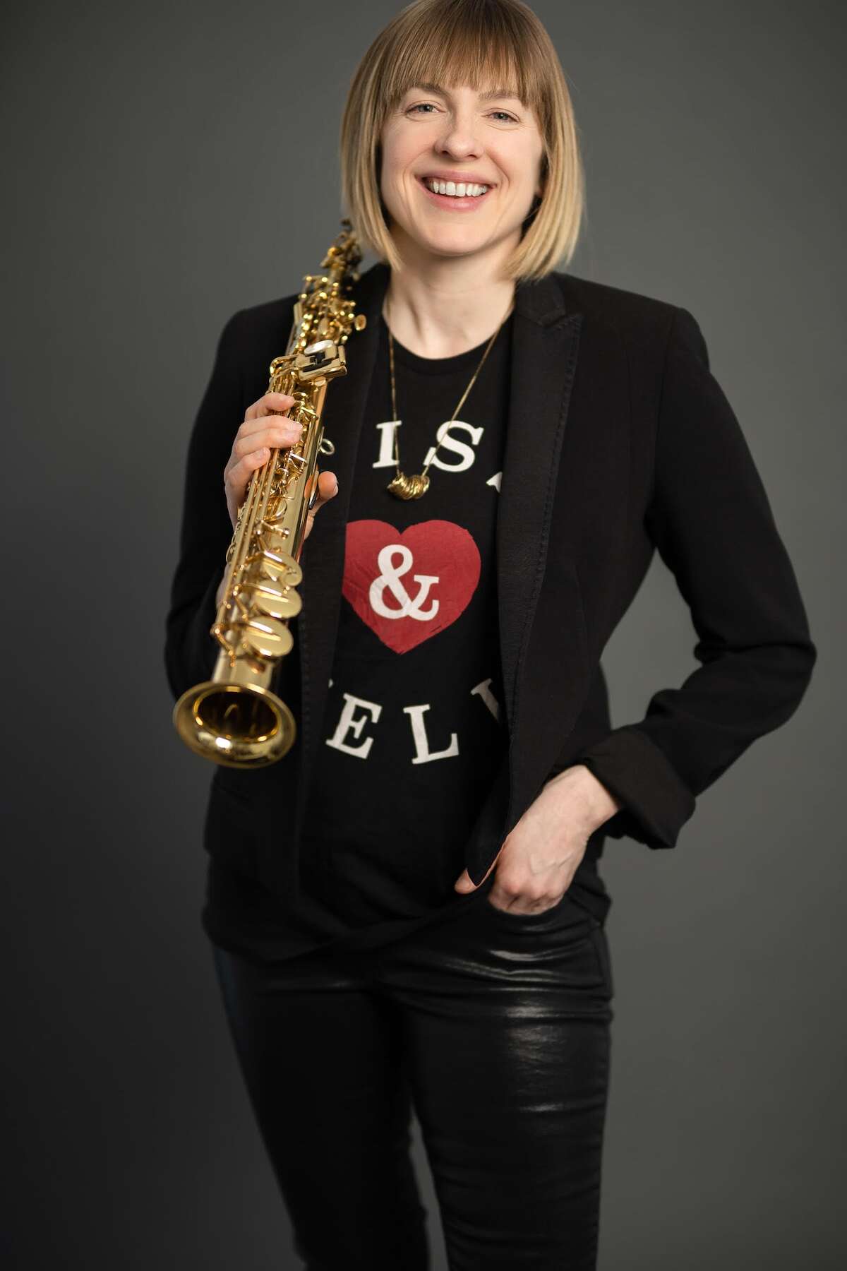 A person with short hair and bangs wearing all black, holding a tenor saxophone in front of a grey background.