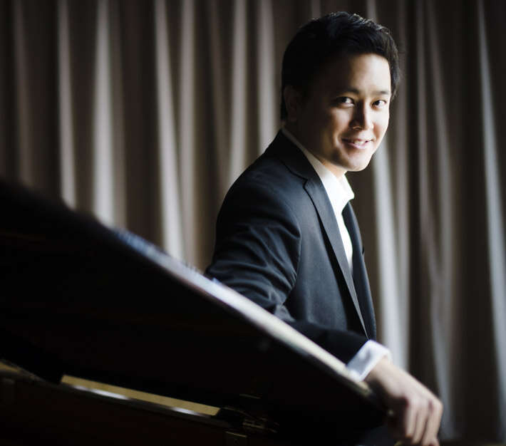 Person wearing a suit, leaning on a grand piano with curtains in the background, smiling.