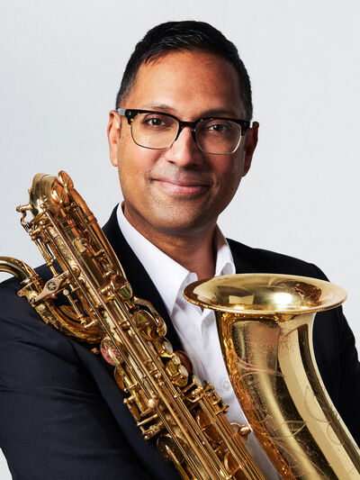 Professional headshot portrait of Shirantha Beddage, smiling and holding his saxophone in front of him against a white backdrop.