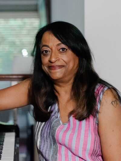 Professional portrait of Professor Bina Jon, sitting down with her arm extended on a piano in front of her inside of a room with a large window.