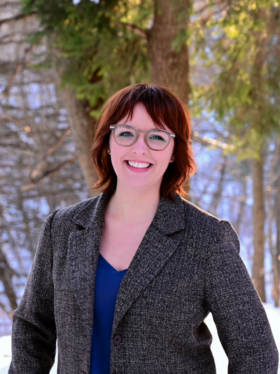 Portrait of Avery Brzobohaty standing outdoors, in front of a wooded area with a grey jacket on