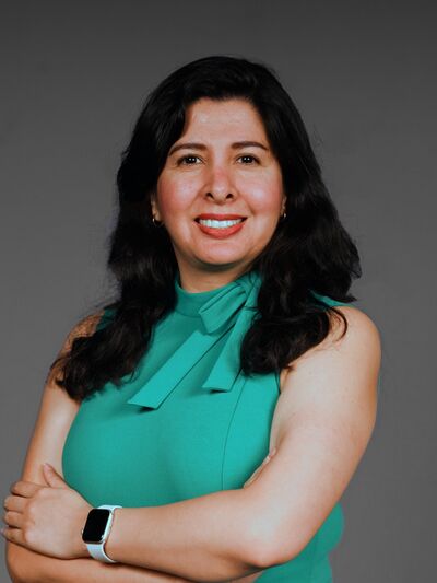 Professional headshot of Cristina Huerta, standing against a dark grey backdrop with her arms crossed and wearing a turquoise top.