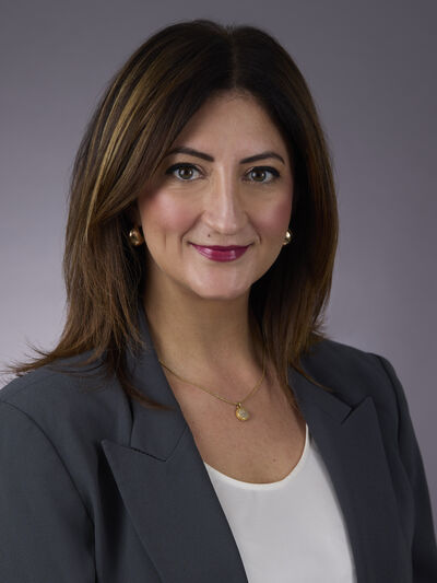 Portrait of woman with brown hair smiling, wearing dark blazer and white top, against a dark grey backdrop