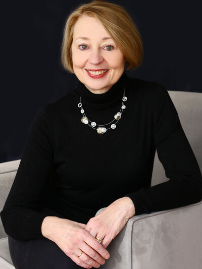 Photo of a woman wearing all black and a silver necklace, posed in a grey chair.