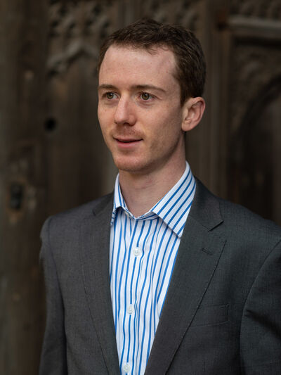 Person with short hair, wearing a shirt and blazer, standing in front of a stone wall with carvings.
