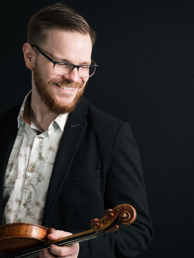 Person smiling, wearing glasses, a button-down shirt, and a blazer, holding a violin.