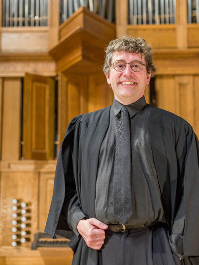 Person wearing glasses and an organist robe, standing in front of an organ.